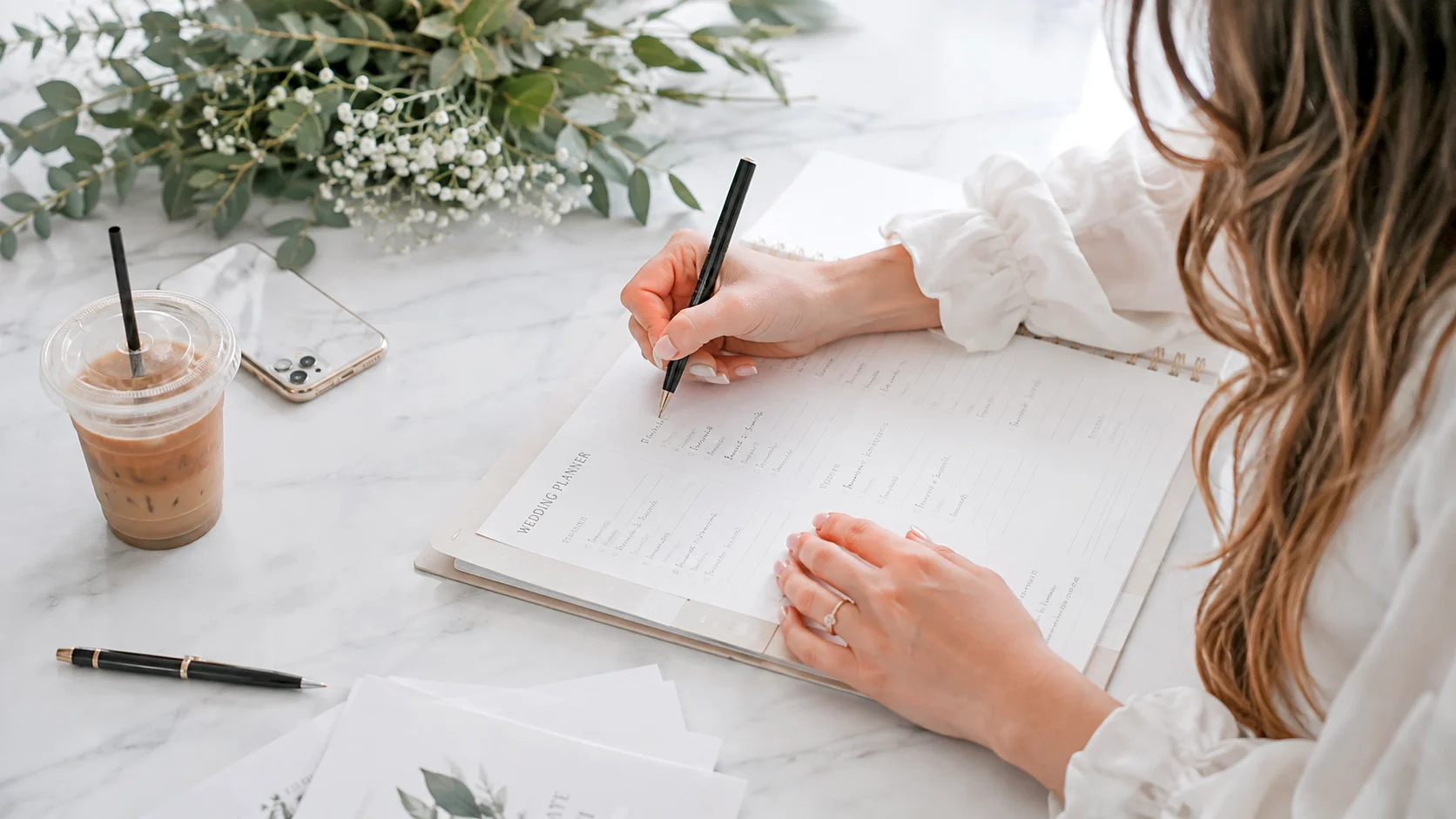 Woman filling out a wedding planner sheet on a white table with greenery and flowers.