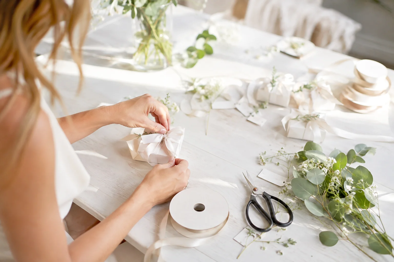 Woman Bride tying pink ribbon onto a white wedding favor box on a white table with greenery and a black small pair of scissors.