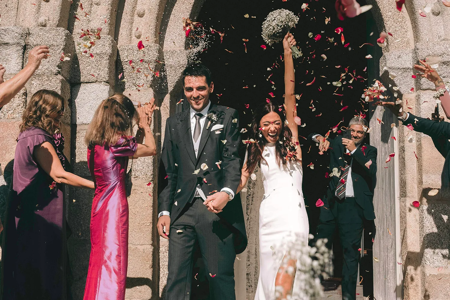 Newlywed bride and groom leaving a church as wedding guests celebrate by tossing colorful flower petals.